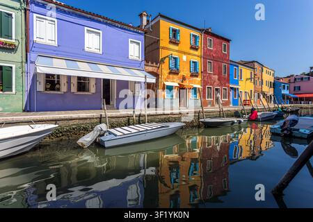 Case colorate sul lungofiume e barche ormeggiate lungo un quieto canale sull'isola di Burano, Italia, con riflessi vividi sotto un cielo estivo sereno. Photographe Foto Stock