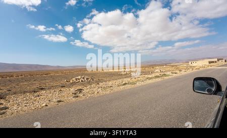 Strada aperta attraverso un paesaggio desertico arido sotto un cielo blu brillante con nuvole bianche, vista dal finestrino di un'auto. Scena di viaggio remota con ampio spazio, roccioso Foto Stock
