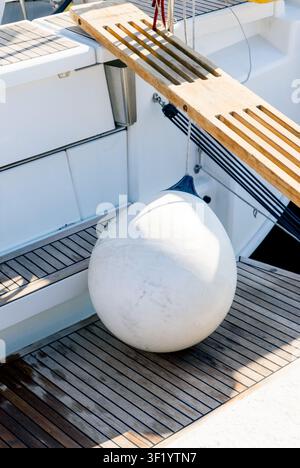 Primo piano di un paraurti bianco per yacht su un ponte in teak accanto a una passerella di legno, evidenziando l'attrezzatura da vela e i dettagli del porto turistico Foto Stock