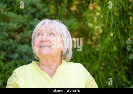 Donna anziana felice che guarda verso l'alto con un sorriso mentre godersi una tranquilla giornata estiva in giardino. Foto Stock