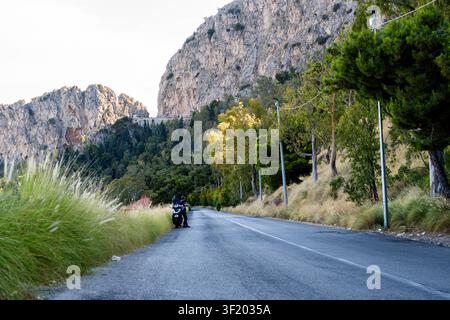Strada panoramica di montagna con uno scooter parcheggiato sul ciglio della strada / Strada asfaltata sotto enormi scogliere in Sicilia Foto Stock