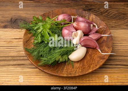 Giovani spicchi d'aglio su piatto di legno, gruppo di spicchi d'aglio, bulbo condimento, ingrediente alimentare naturale con prezzemolo verde e aneto su tavolo di legno rustico Foto Stock