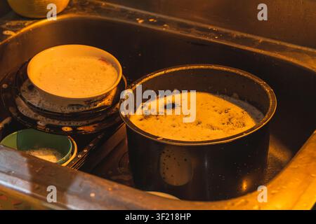Un lavello da cucina in acciaio inossidabile pieno di acqua saponata, ciotole sporche, piatti e una grande pentola nera che si scioglie prima di essere lavata. Foto Stock
