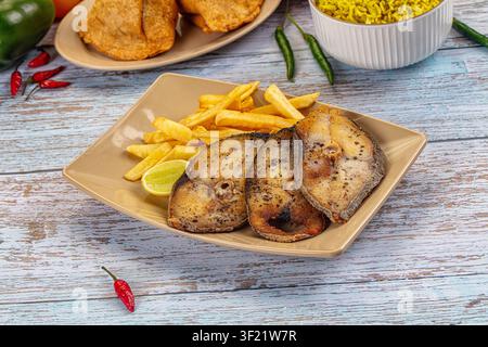 Tre bistecche di pesce grigliate con pelle croccante servite con patatine dorate e una fetta di lime su un piatto beige. Tavolo rustico in legno blu con peperoncini rossi e verdi, giallo r Foto Stock