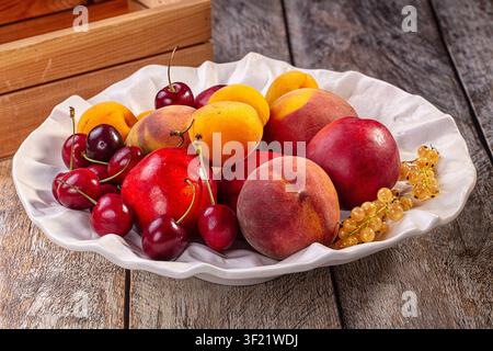 Ciliegie, pesche, albicocche, ribes bianchi vibranti disposti in una ciotola bianca a forma di conchiglia. Una luce naturale calda evidenzia le texture succose dei rossi intensi, giallo Foto Stock