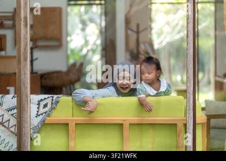 Una donna indonesiana musulmana asiatica in hijab con il suo bambino appoggiato a un divano verde, vista attraverso una finestra di vetro Foto Stock