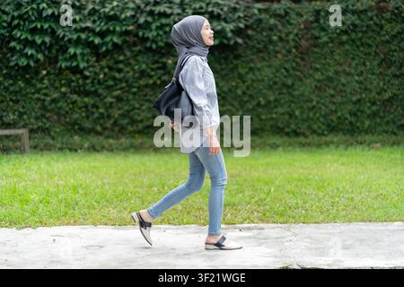 Giovane donna musulmana indonesiana asiatica in abbigliamento casual e hijab che cammina all'aperto, portando una borsa a mano con un sorriso Foto Stock