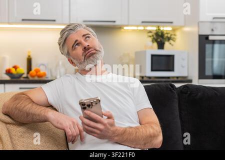 Uomo di mezza età a casa scorre le notizie sullo smartphone si sente annoiato, stanco e stressato e fa un sospiro silenzioso. Uomo maturo sul divano abbassa il telefono, si appoggia indietro, umore a bassa energia, occhi e mente rallentano e hanno bisogno di una pausa. Foto Stock