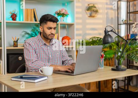 Un uomo indiano seduto al tavolo di casa lavora su un laptop, beve caffè, sorride e ritorna al suo compito di lavoro remoto concentrato. Un ragazzo arabo seduto al tavolo di casa si gode una bevanda calda durante il lavoro online in un soggiorno accogliente con un'atmosfera tranquilla. Foto Stock