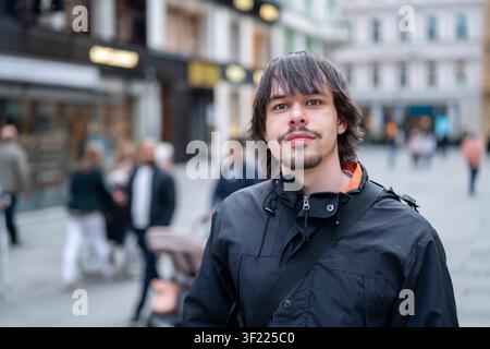 Ritratto di un giovane uomo in piedi all'aperto su una strada pedonale urbana con persone ed edifici sfocati sullo sfondo. Indossa una giacca scura Foto Stock