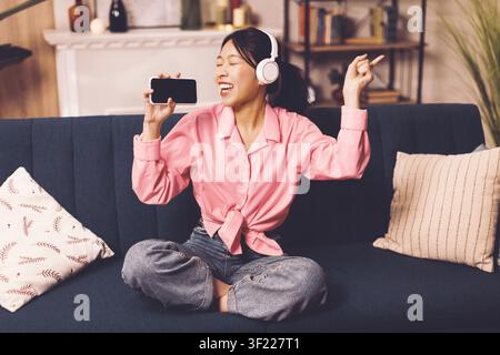 Donna asiatica felice che ascolta musica con cuffie e smartphone a casa, rilassandosi Foto Stock
