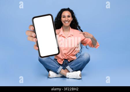 Donna seduta sul pavimento che tiene uno smartphone e indica lo schermo in un ambiente da studio Foto Stock