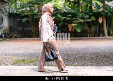 Donna musulmana indonesiana in hijab che cammina all'aperto portando un tablet e una borsa a mano, rappresentando uno stile di vita professionale moderno, mobilità e collaborazione professionale Foto Stock