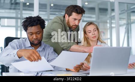Tre partner aziendali multirazziali concentrati, squadra formale che gestisce documenti con laptop, servizio aziendale in ufficio, colleghi multietnici Foto Stock