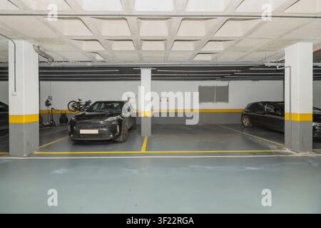 Garage sotterraneo con pavimento grigio, linee gialle, pilastri bianchi, due auto nere e biciclette montate a parete. Foto Stock