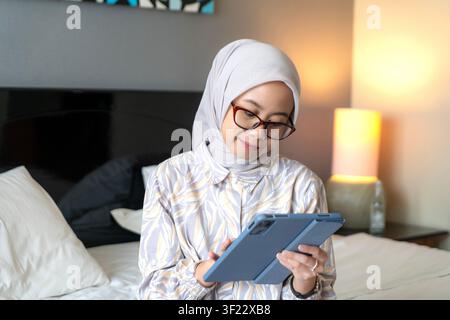Donna indonesiana asiatica con hijab e occhiali, che sorride mentre tiene un tablet e si siede su un letto al chiuso. Foto Stock