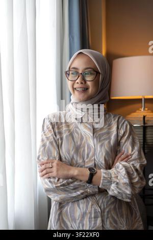 Ritratto di una donna musulmana indonesiana asiatica con hijab e occhiali, che guarda fuori da una finestra luminosa all'interno con un sorriso. Foto Stock