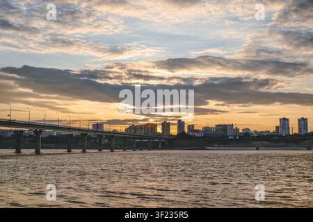 Volgograd, Russia - 25.05.2023: Silhouette della città con ponte sul fiume Volga durante il tramonto. Cielo drammatico con nuvole e luce calda della sera che si riflette Foto Stock