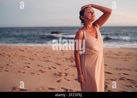 Ritratto di una donna pacifica in piedi sulla spiaggia sabbiosa di Tangalle durante un caldo tramonto, indossando un vestito elegante, rilassandosi e godendosi la vista calma dell'oceano con dolce Foto Stock