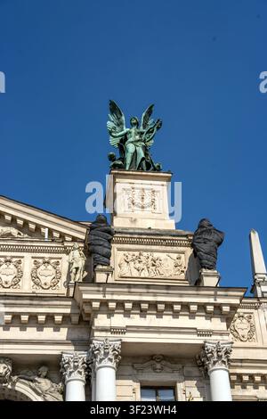 Una statua di angelo si erge sulla sommità di un edificio storico. Due figure sono visibili ai lati della statua. L'ambientazione ha un cielo blu chiaro e luminoso Foto Stock