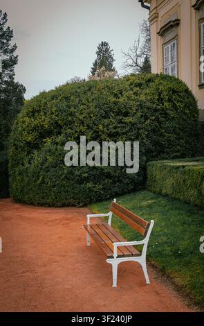 Uno scatto verticale e sereno di una panchina di legno vuota posta su un sentiero di ghiaia rossa in un tranquillo parco del castello. Foto Stock