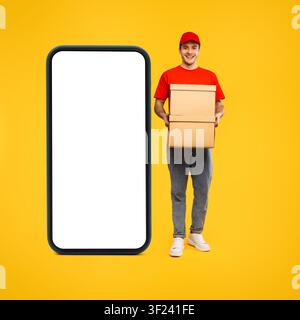 Giovane uomo caucasico felice che tiene scatole di cartone, modello di smartphone su sfondo giallo Foto Stock