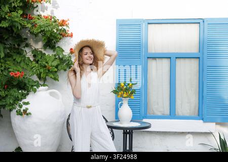 Una donna allegra che indossa un cappello di paglia posa in un ambiente esterno luminoso e soleggiato con fiori vivaci e un tavolo adornato di limoni freschi. Foto Stock