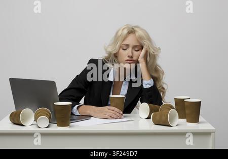 Giovane donna caucasica preoccupata in abito con tazze di caffè alla scrivania, concetto di sovraccarico lavorativo Foto Stock