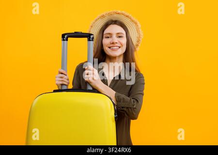 Giovane donna sorridente che tiene una valigia gialla con cappello di paglia, concetto di viaggio Foto Stock