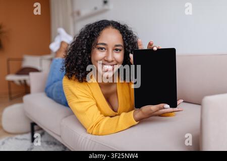 Donna nera sorridente che mostra un tablet in un soggiorno accogliente, concetto di stile di vita moderno Foto Stock