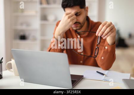 Uomo mediorientale preoccupato che tiene occhiali, stressato al computer portatile in un ufficio moderno, difficoltà lavorative Foto Stock