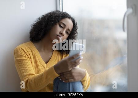 Giovane donna nera pensierosa che usa lo smartphone seduta vicino alla finestra, stato riflessivo Foto Stock