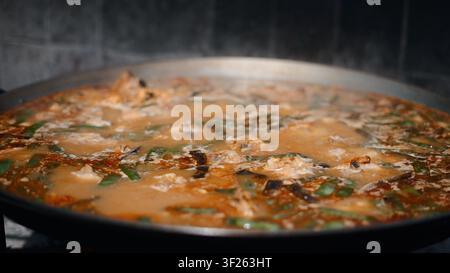 Paella tradizionale che cuoce lentamente in una grande padella con vapore e ingredienti Foto Stock