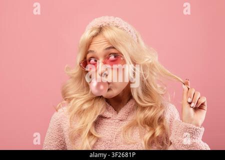 Giovane donna che morde una gomma da masticare e gioca con i capelli, sfondo rosa, atmosfera gioiosa Foto Stock