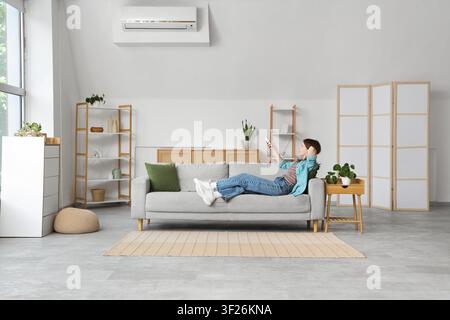 Giovane donna che si rilassa su un comodo divano regolando l'aria condizionata con un telecomando in un divisorio moderno del soggiorno Foto Stock