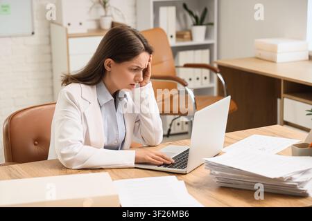 Giovane donna d'affari frustrata che sperimenta stress e burnout mentre lavora su un laptop in un ambiente d'ufficio moderno, tiene la mano alla testa Foto Stock