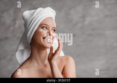 Giovane donna che sorride mentre applica una crema per la cura della pelle a casa, concetto di bellezza Foto Stock