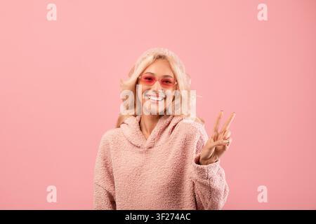 Giovane donna sorridente con capelli biondi e occhiali rosa che fa il segno della pace su sfondo rosa Foto Stock