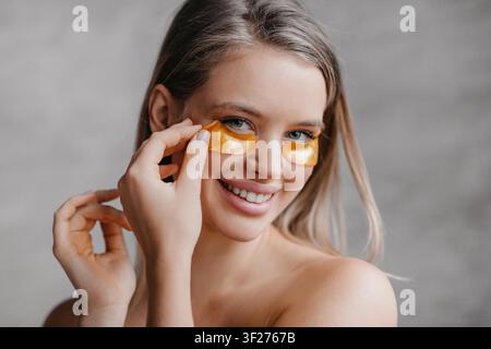 Giovane donna sorridente con maschere per gli occhi in oro che applica trattamenti di cura a casa, concetto di benessere Foto Stock