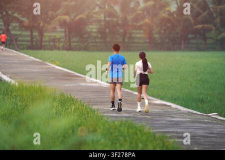 Vista posteriore di un giovane uomo e una giovane donna asiatici che fanno jogging su un sentiero rurale circondato da campi verdeggianti. Stile di vita sano, esercizio all'aperto e fitness mattutina Foto Stock
