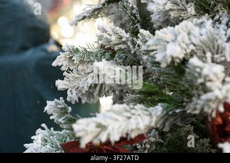 Rami sempreverdi coperti di neve riempiono una vetrina del mercato natalizio, con decorazioni rosse parziali e luci sfocate visibili intorno al bianco denso Foto Stock