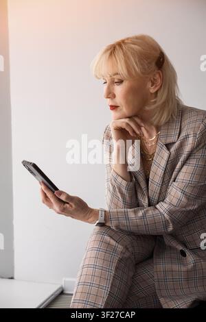 Donna d'affari bionda anziana in un completo a quadri è seduta al chiuso, concentrata sul suo smartphone, sfogliando pensierosa contenuti digitali ed email, trasmette Foto Stock