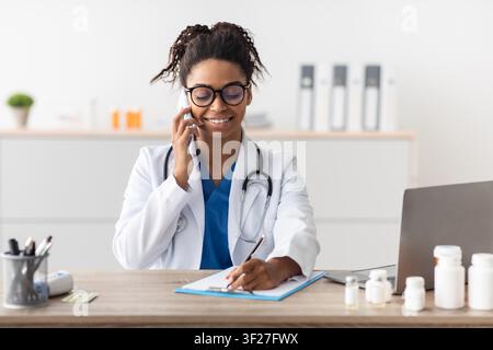 Donna medico nera felice che fa una telefonata e prende appunti nel suo ufficio, concetto di assistenza sanitaria Foto Stock