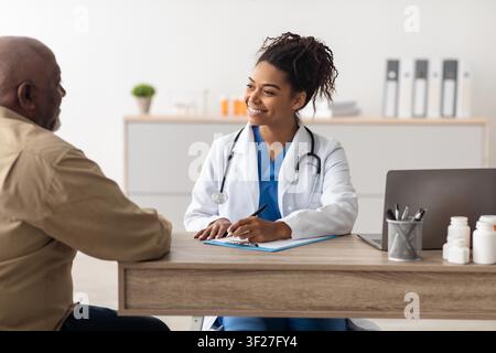 Dottoressa nera sorridente che consulta un paziente maschio anziano in una clinica moderna, concetto di assistenza sanitaria Foto Stock