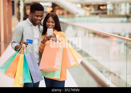 Coppia nera felice che fa shopping insieme al centro commerciale, guarda lo smartphone con le borse della spesa Foto Stock