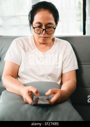 Donna asiatica di mezza età con occhiali che usa lo smartphone sul divano a casa, concetto di banking online, app mobile, comunicazione digitale, telemedicina, un Foto Stock