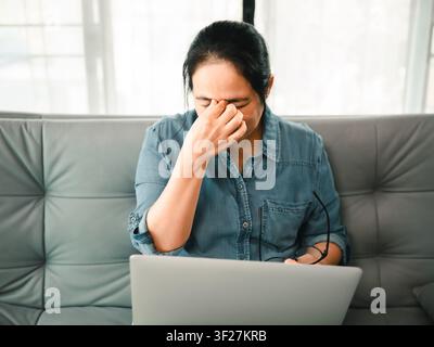 Donna asiatica stanca che si strofina gli occhi mentre è seduta con un laptop e gli occhiali a casa, concetto di affaticamento oculare, stanchezza digitale, mal di testa, tempo sullo schermo, sovraccarico, Foto Stock