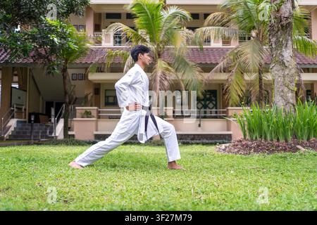 Uomo indonesiano asiatico che esegue una posizione avanti in karate durante l'allenamento di arti marziali all'aperto su un campo erboso con uno sfondo di edificio tropicale. Foto Stock