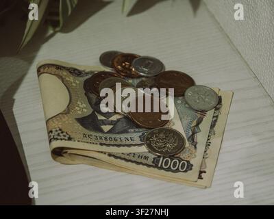 Banconote giapponesi da 1000 yen piegate con varie monete da yen impilate su un tavolo di legno bianco Foto Stock