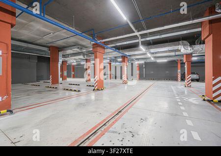 ampio garage sotterraneo con file di pilastri arancioni, tubi blu visibili e auto parcheggiate in lontananza Foto Stock